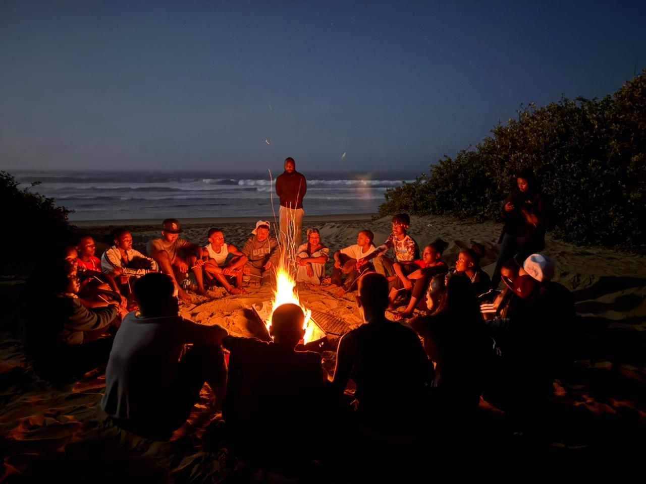 high school students sitting around a camp fire at the beach at night